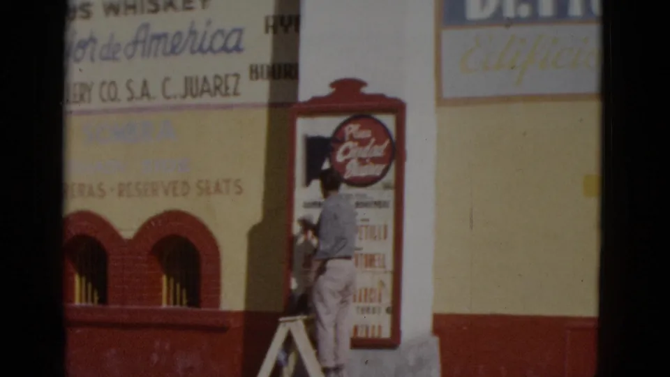 Continued shot of worker on ladder at a building exterior displaying arena and commercial signage, likely a bullring facade in northern Mexico, c.1952.