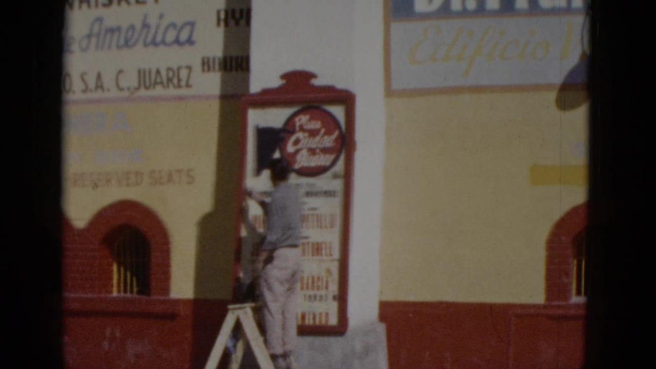 Man on a wooden ladder adjusting a circular venue sign on a yellow and red building facade.