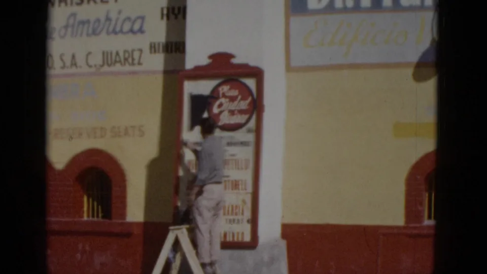 Man on wooden ladder tending a 'Plaza Ciudad Juarez' circular sign on a yellow terracotta building; northern Mexico, circa 1952, 8mm film.