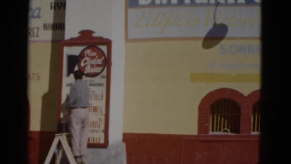 Worker on ladder at a yellow and terracotta building with 'Plaza Ciudad Juarez' sign and 'Edificio Victoria' text; northern Mexico, circa 1952.