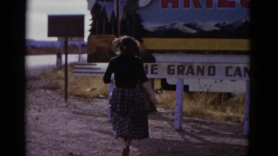 Woman in a dark top and plaid skirt walks beside a roadside sign in Baja California, Mexico, with desert landscape beyond.