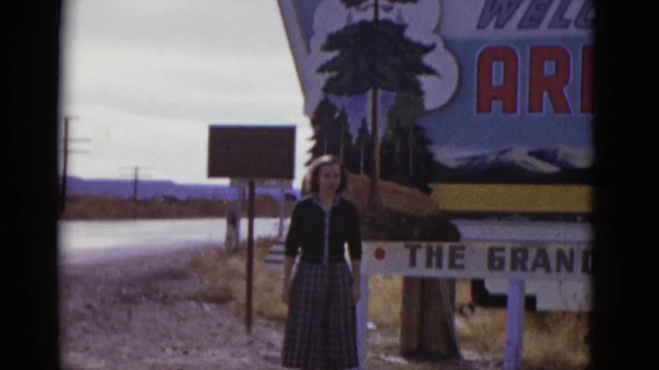 Woman in a plaid skirt stands in front of a large travel sign with visible Arizona and Grand Canyon lettering, on 1952 8mm footage.