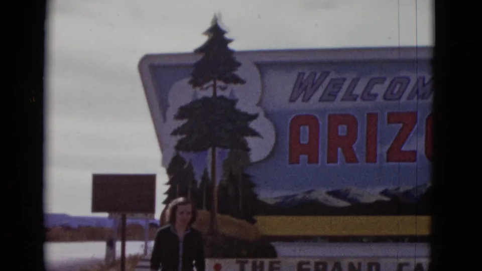 The woman remains near the lower edge of the Welcome to Arizona sign, with the sign lettering and painted landscape filling most of the frame.