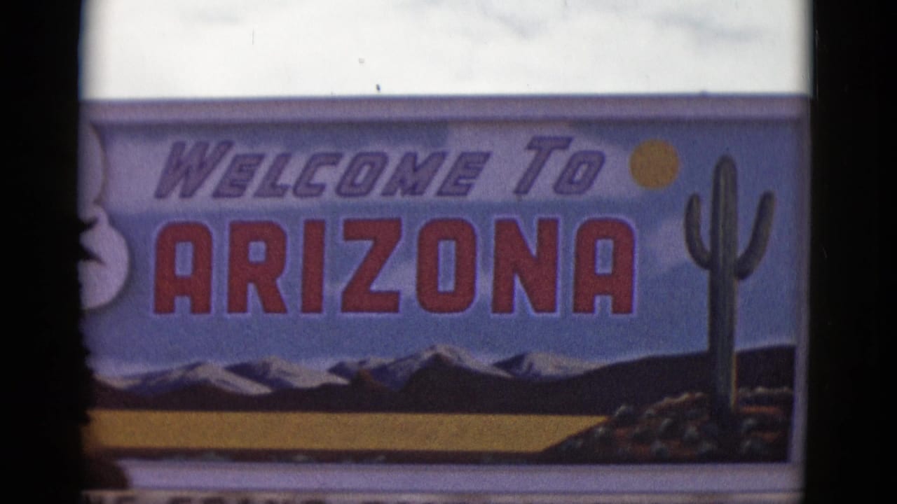 1952 Arizona home-movie frame showing a Welcome to Arizona roadside sign with red lettering, cactus artwork, a sun symbol, and illustrated mountains.