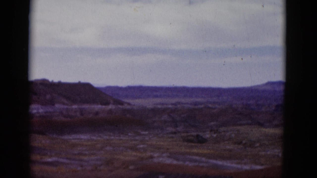 Wide desert valley with low hills, distant mesa, pale sky, and dark film borders.