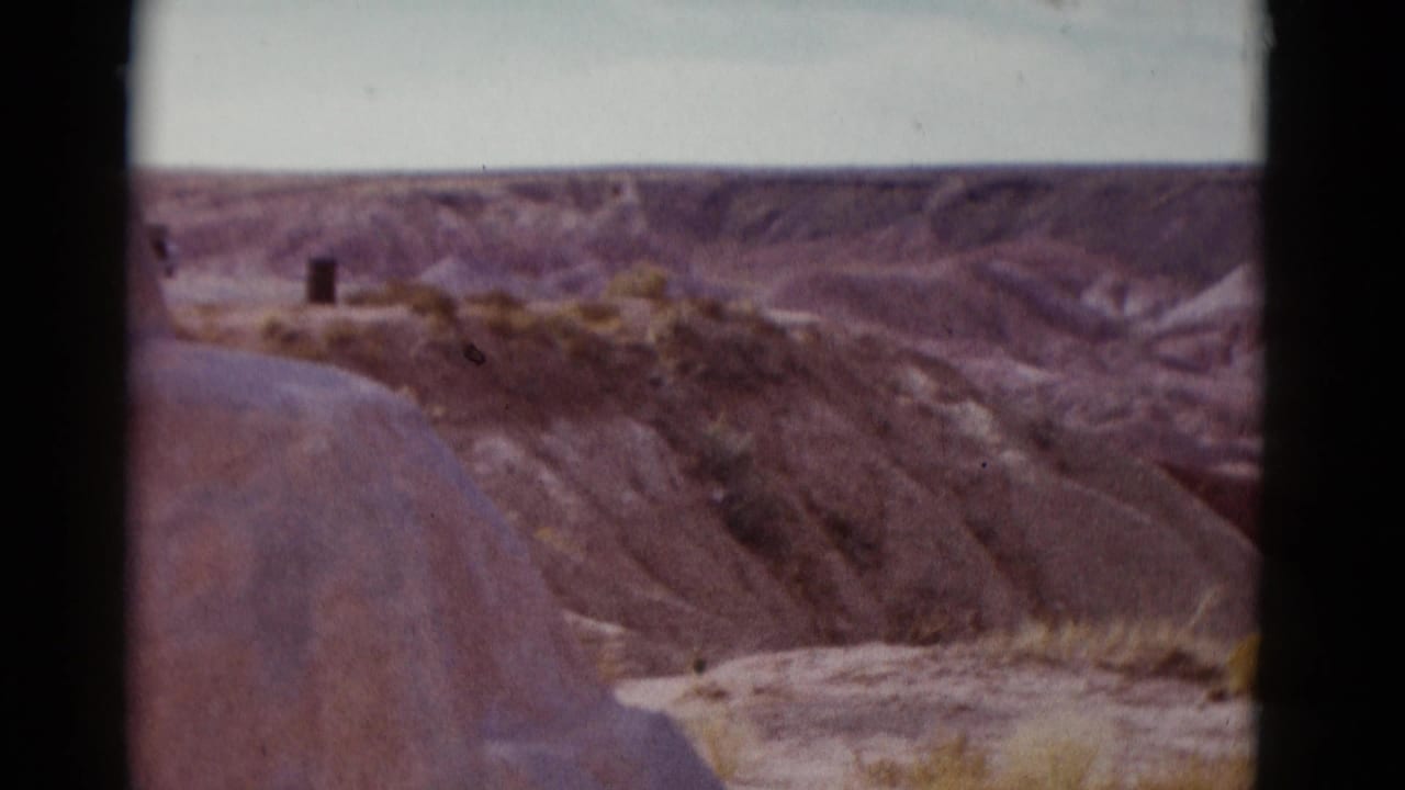 Dry eroded desert hills under a pale sky with sparse shrubs and dark 8mm frame edges.