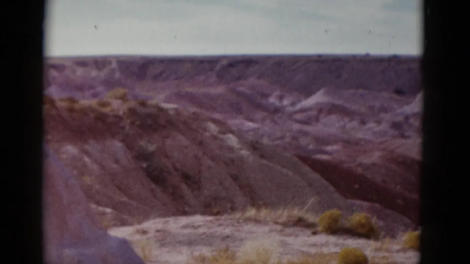 A mid-century 8mm travel shot holds on Arizona desert hills, dry wash-like cuts, and sparse foreground shrubs.