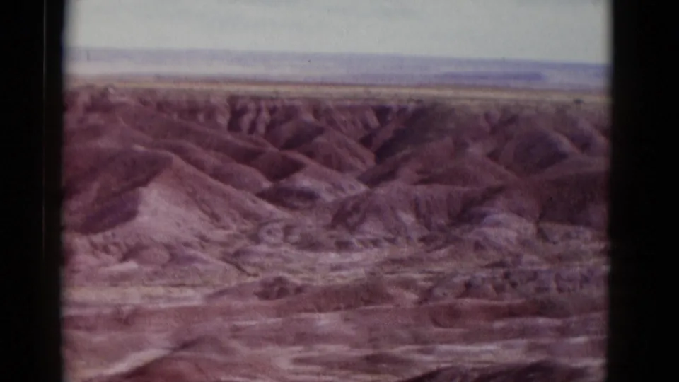 1952 Arizona home-movie frame with rolling desert badlands, layered washes, muted pink-brown hills, and a pale horizon in soft 8mm color.