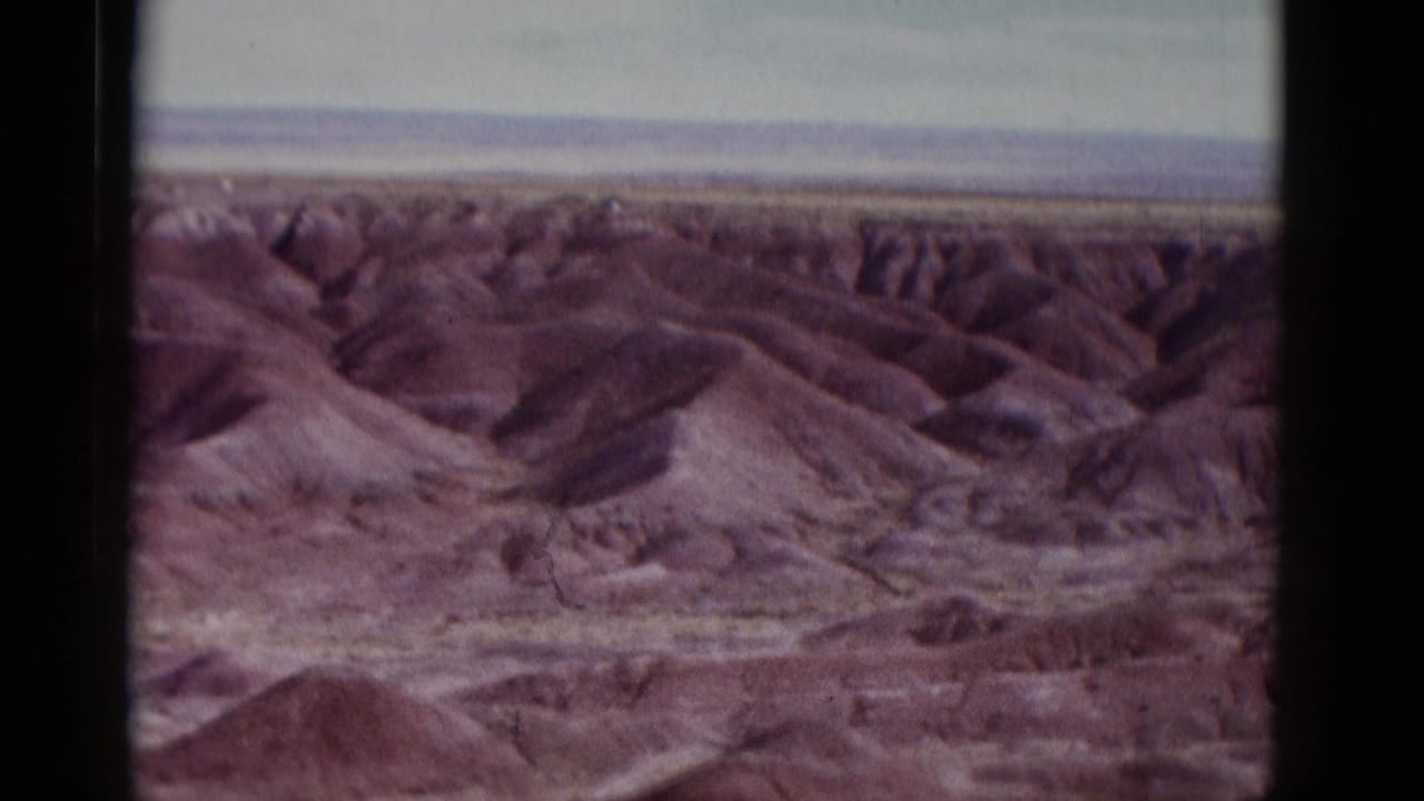 Reddish eroded desert hills beneath a pale horizon, framed by dark film borders.