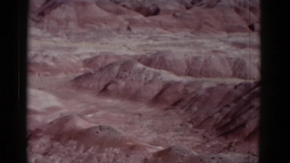 Faded 8mm frame of rugged Arizona desert ridges and eroded washes, with pale sky and visible scan scratches.
