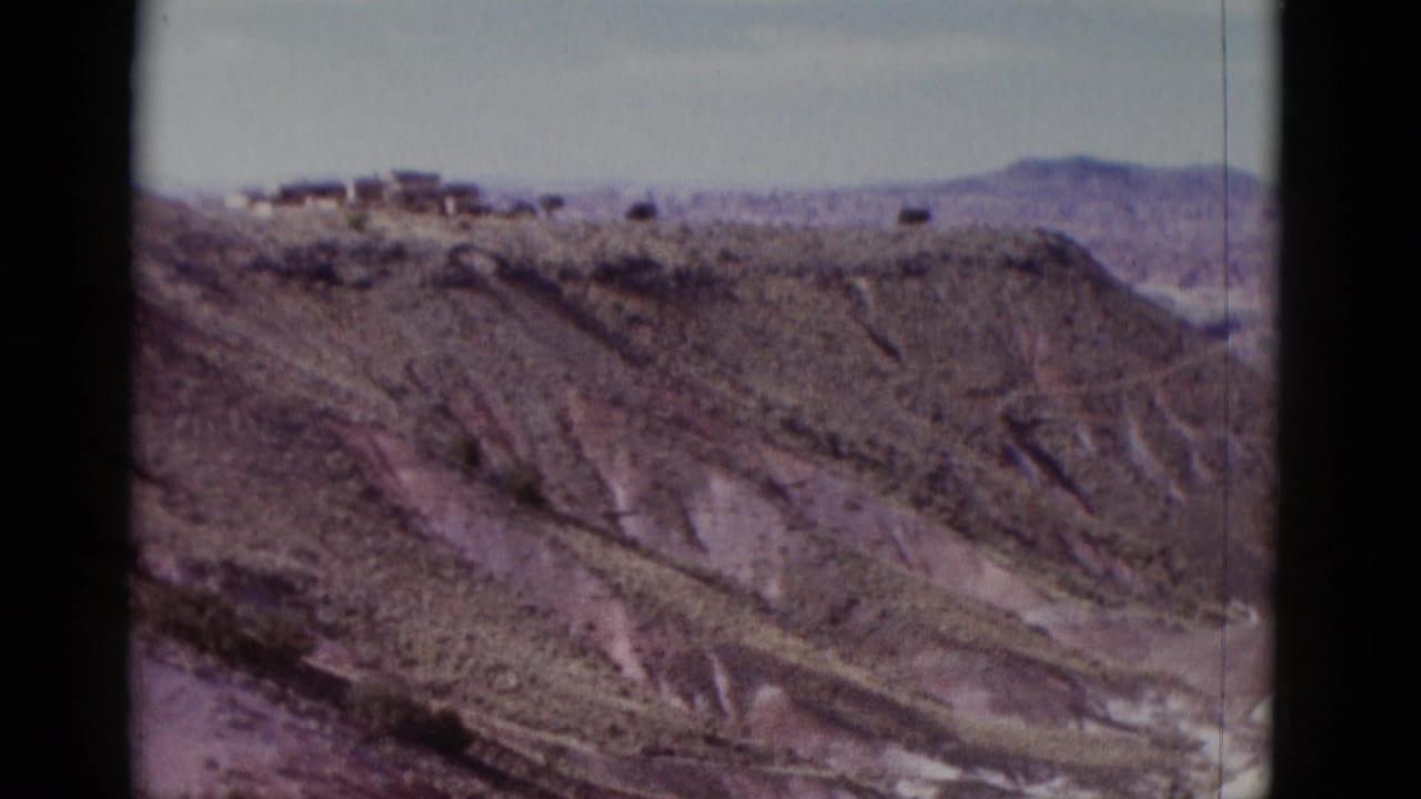 1952 Arizona travel footage frame with eroded desert slopes, distant mountains, and small ridge-top structures.