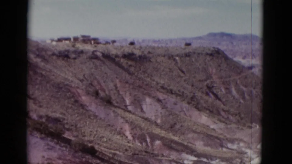 1952 Arizona travel footage frame with eroded desert slopes, distant mountains, and small ridge-top structures.
