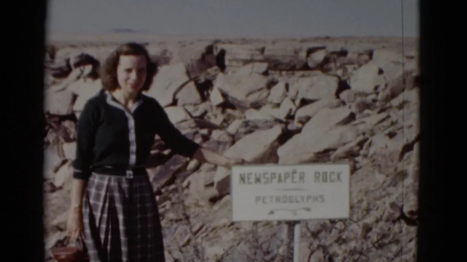 In 1952 Arizona, a woman in a dark cardigan and plaid skirt poses beside the Newspaper Rock Petroglyphs sign among pale boulders.