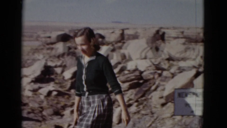 1952 Arizona home-movie frame of the woman turning near boulders, with part of the Newspaper Rock sign visible at the right edge.