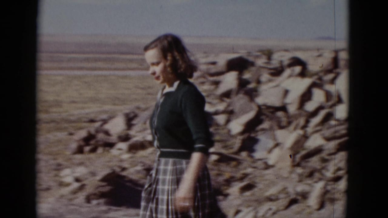 Young woman in a dark cardigan and plaid skirt walks through rocky Arizona desert terrain in 1952 home-movie footage.