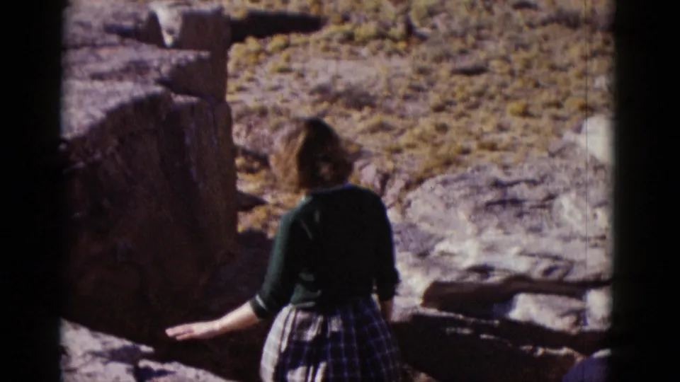 Woman in a dark cardigan and plaid skirt reaches toward a large rock in the Arizona desert landscape.