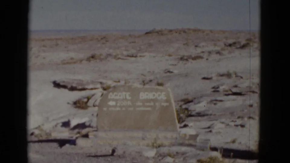 1952 Arizona frame showing an Agate Bridge stone marker with a directional arrow against open desert and pale sky.