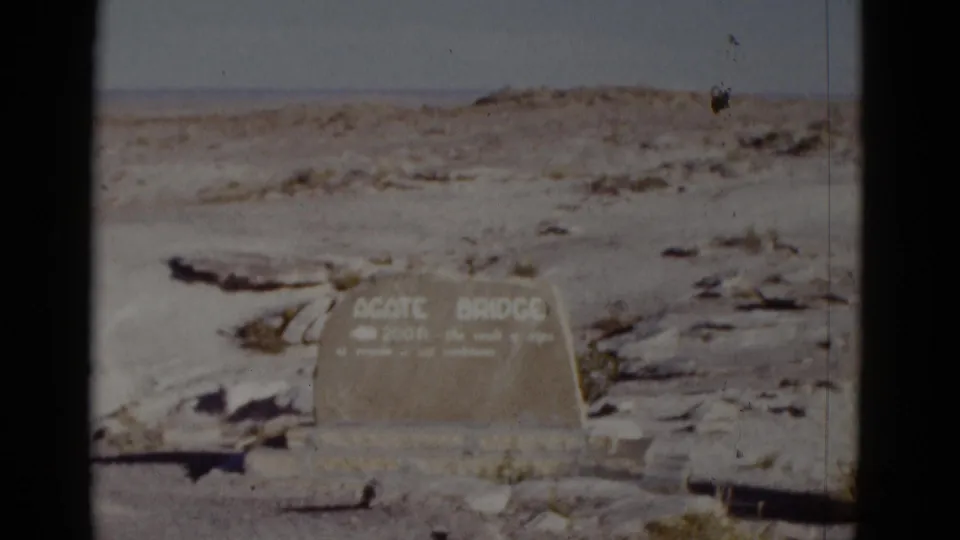 1952 Arizona home-movie frame showing a weathered Agate Bridge sign in pale rocky desert ground under a faded sky.