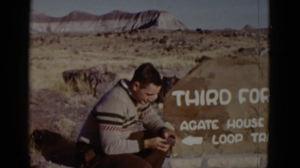 1952 Arizona home-movie frame showing a young man in a striped sweater seated beside a Third Forest and Agate House Trail sign.