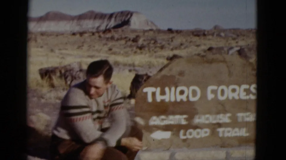 1952 Arizona frame showing a young man sitting next to the Third Forest and Agate House Trail sign with a mesa in the distance.