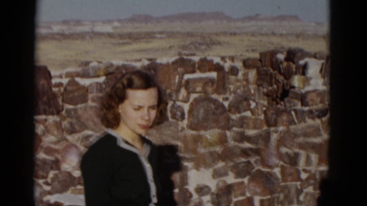 Woman in a dark dress stands beside a rough stone wall in desert light.