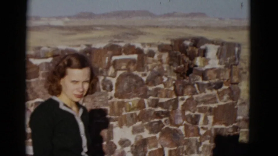 Closer 1952 Arizona home-movie view of the woman near the weathered wall, with desert terrain and pale sky behind her.
