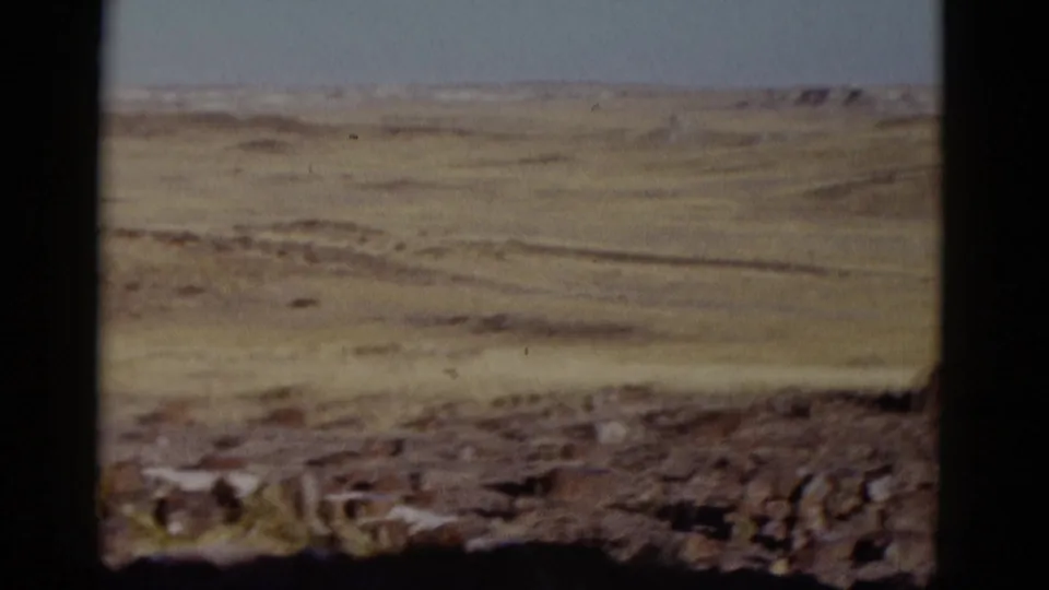 1952 8mm Arizona travel frame showing open grassland, rough rocks in the foreground, distant hills, and dark gate borders.