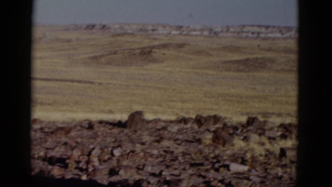 Wide arid plain beyond rocky ground, with low ridges on the horizon and dark frame borders.