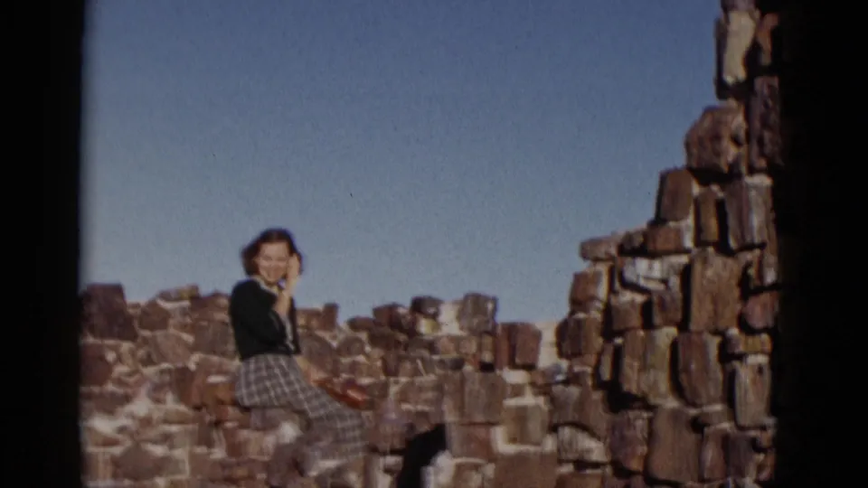 Arizona 1952 home-movie frame showing a smiling woman in a dark sweater and plaid skirt posing on rugged stonework.