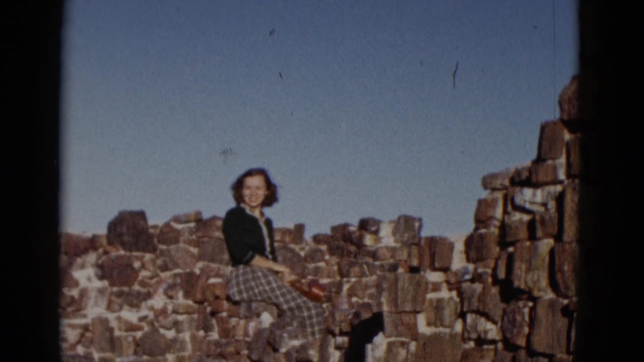 In Arizona home-movie footage from 1952, a woman in a dark sweater and plaid skirt sits on weathered stone ruins.