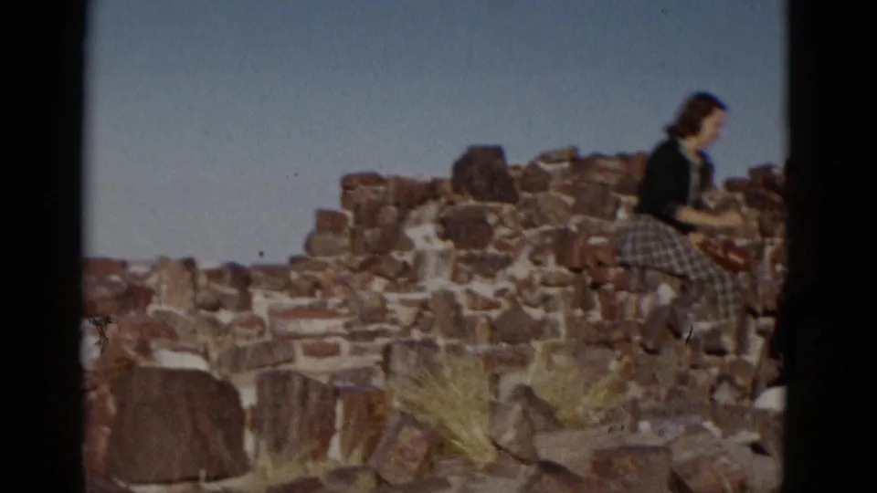 The camera catches the woman in profile on the ruin wall, with uneven stones and desert grasses below.