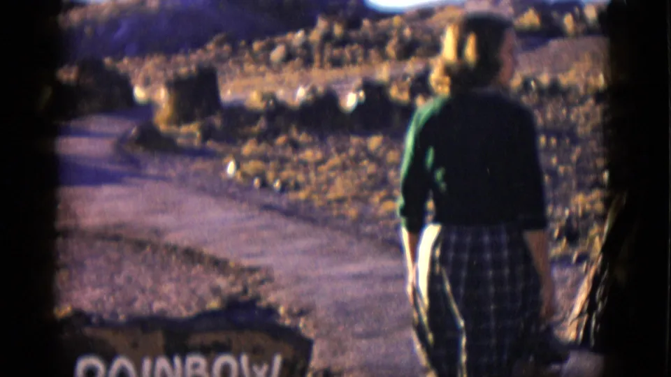 1952 Arizona home-movie frame of a woman in a green top and plaid skirt walking away along the path near the Rainbow Forest sign.