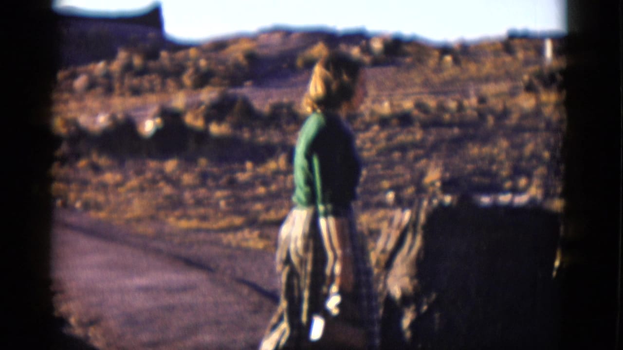 Woman in a green sweater stands on a dirt path in a rocky desert landscape.
