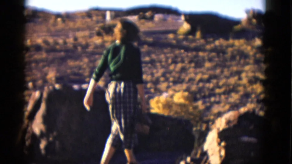 The woman walks through a rocky Arizona desert landscape, with sparse vegetation and low hills in the background.