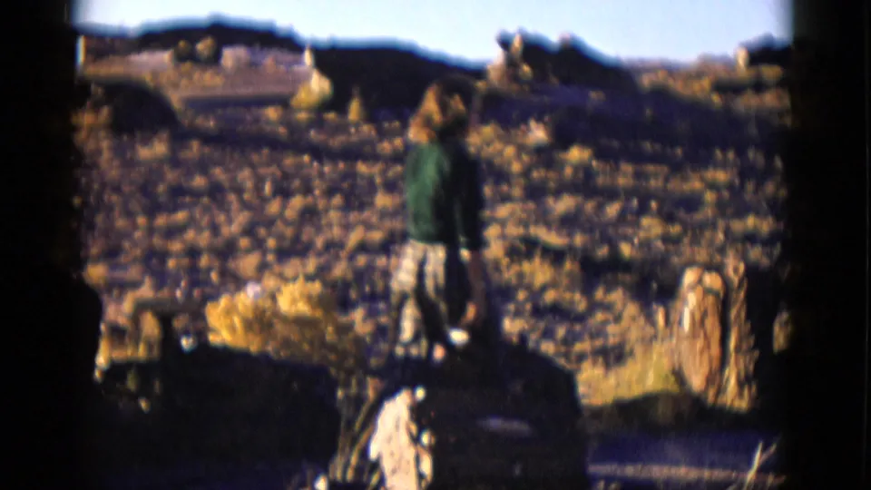A home-movie frame shows the woman amid desert rocks and brush, facing away across an Arizona landscape.