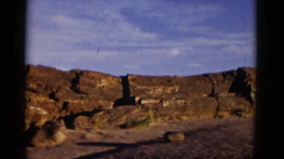 1952 Arizona 8mm frame showing a low line of brown stones, sparse desert floor, and clouds in a blue sky.