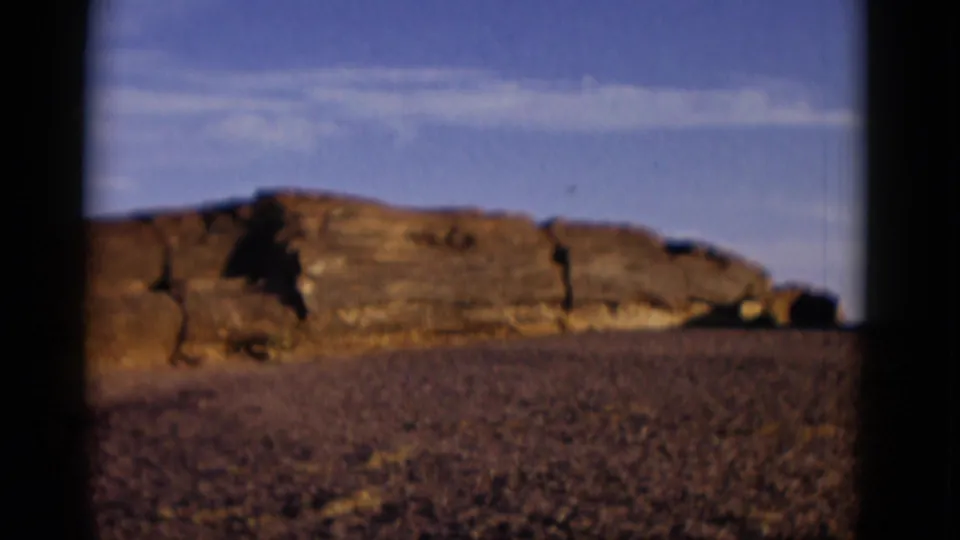 1952 Arizona 8mm view of a layered rock ledge crossing the frame with gravelly desert foreground and blue sky above.
