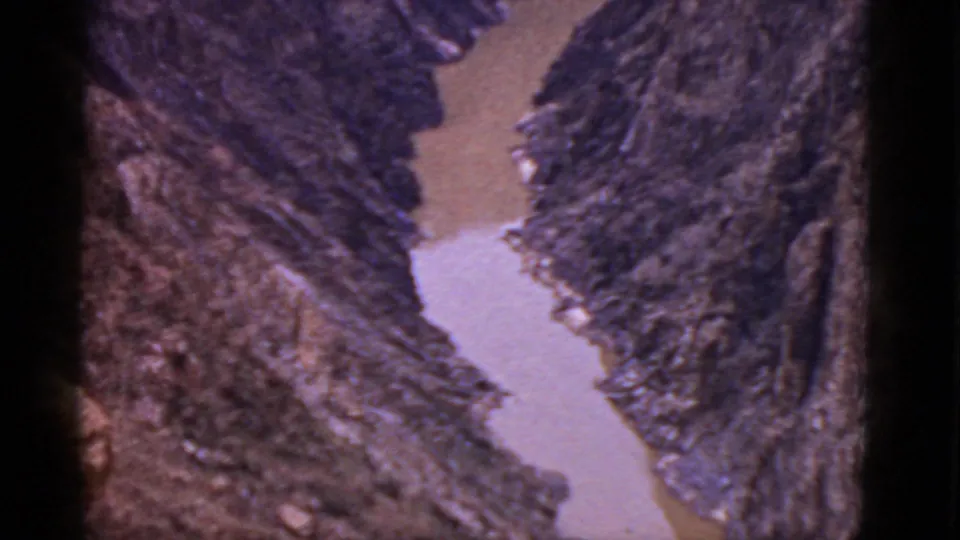 1950s home-movie frame of a muddy Arizona river channel running through a rugged mountain valley.