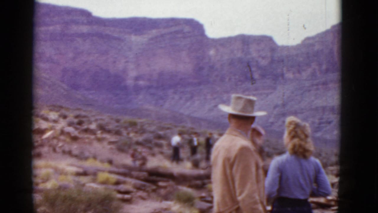 In metadata-supported 1952 Arizona home-movie footage, a man in a light cowboy hat and a woman in a blue top stand with their backs to the camera facing mountains and distant riders.