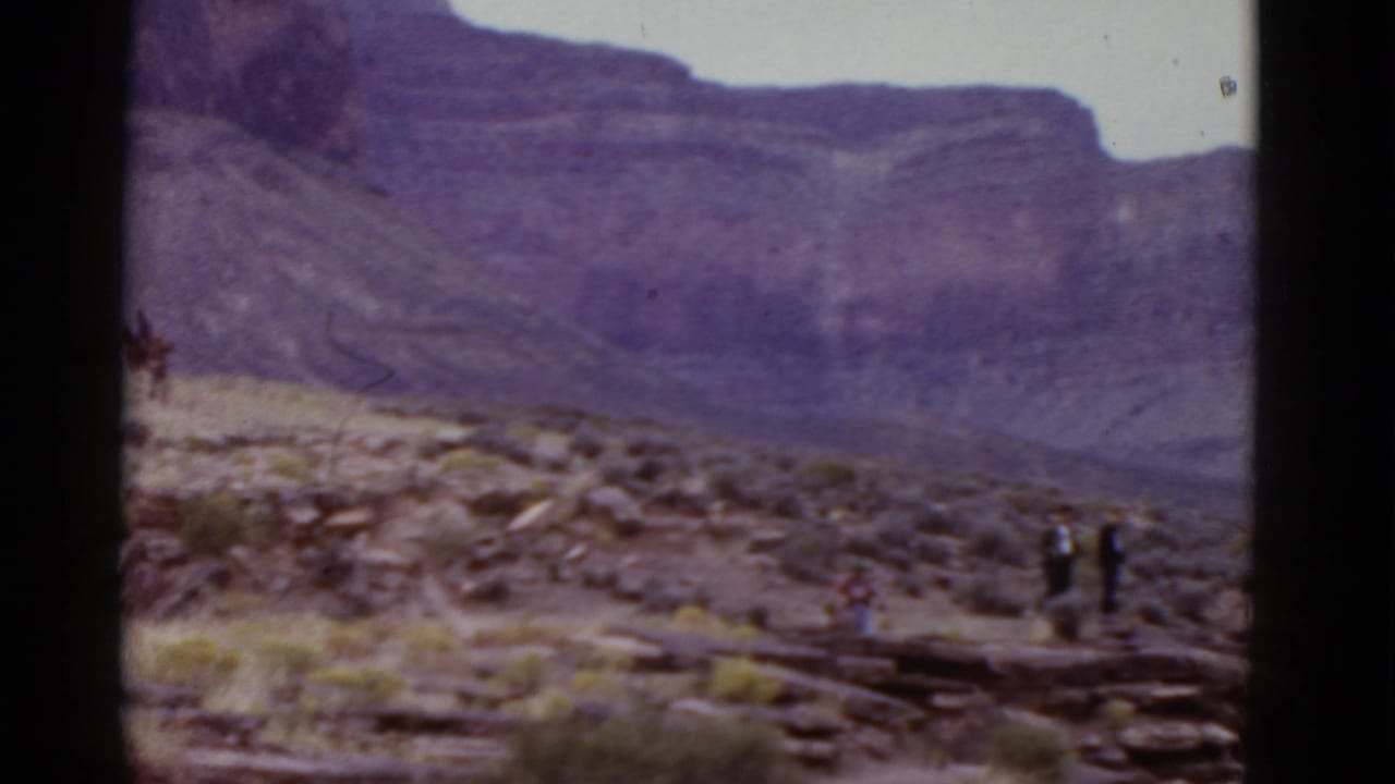 A wide 8mm view shows a rocky Arizona desert slope with small distant figures near a trail and a large purple-hued mountain wall in the background.