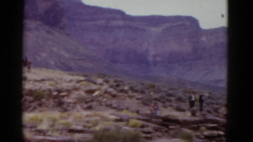 A wide 8mm view shows a rocky Arizona desert slope with small distant figures near a trail and a large purple-hued mountain wall in the background.