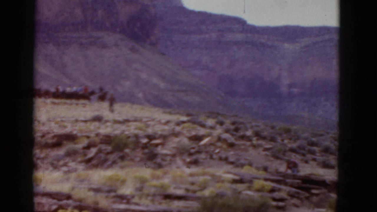 Line of distant riders or people moves across rocky desert terrain beneath mountains.