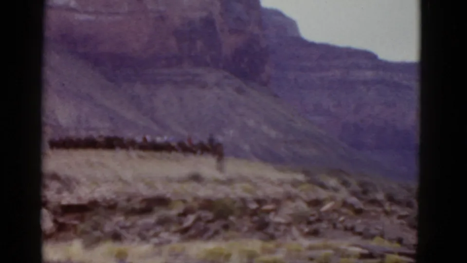 A wide, soft-focus view shows a distant group, likely horseback riders, moving along a desert ridge against a purple-toned Arizona mountain backdrop.