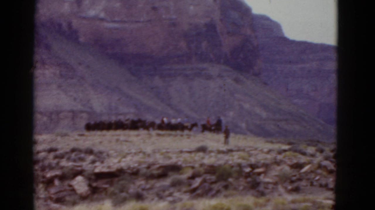 Long line of horses and riders crossing rocky desert below layered cliffs.