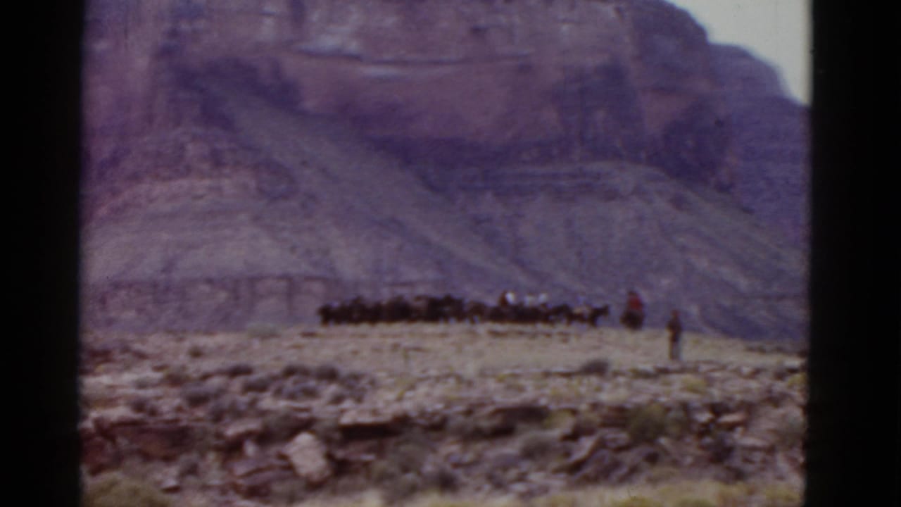 Distant riders and horses travel across desert terrain with mountains behind them.