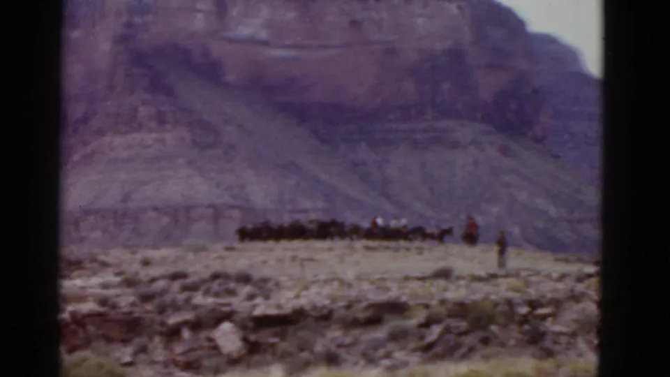 1952 Arizona frame with a long group of horses and riders moving across pale desert before layered mountain cliffs.