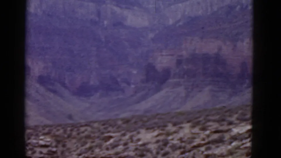 1952 Arizona home-movie frame showing a layered cliff face above rocky scrub terrain with a purple aged-film cast.
