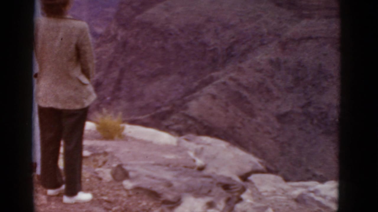 1952 Arizona travel frame with a woman in a light jacket and dark pants facing a canyon from a rocky overlook.