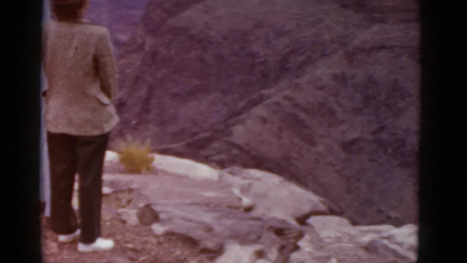 1952 Arizona travel frame with a woman in a light jacket and dark pants facing a canyon from a rocky overlook.