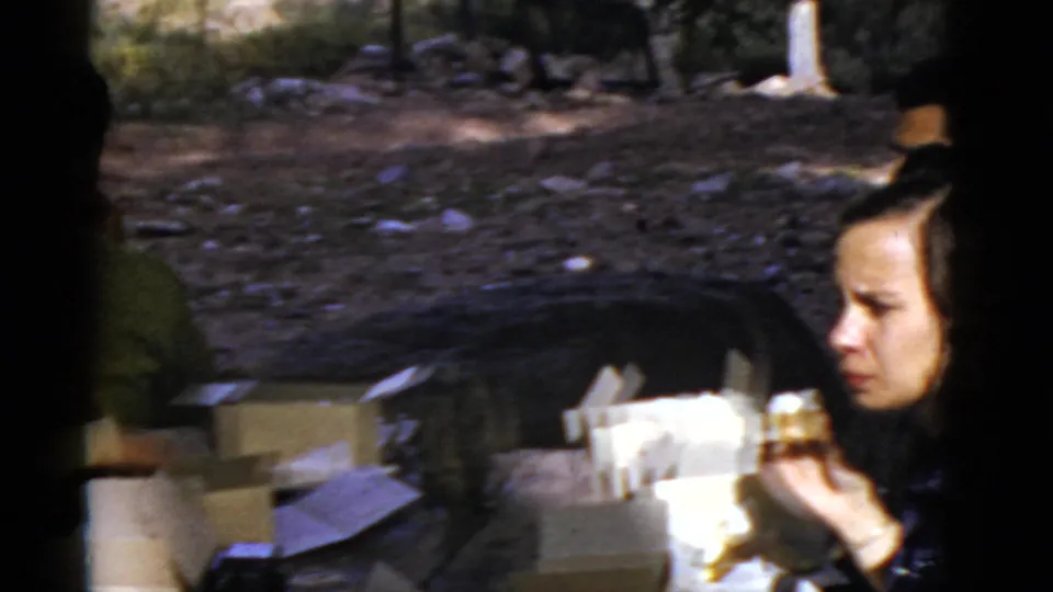 1952 Arizona home-movie frame showing a woman holding food beside picnic boxes on rocky ground.